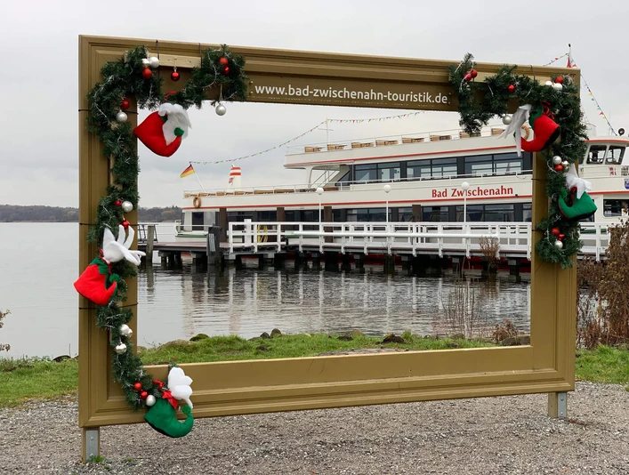 winter-wunder-welt-2021-bilderrahmen.jpg Weihnachtlich geschmückter Bilderrahmen mit Blick auf das Fahrgastschiff Bad Zwischenahn am See.