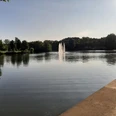Ruhiger Weiher mit Springbrunnen, umgeben von grünen Bäumen und Promenade, unter klarem Himmel.