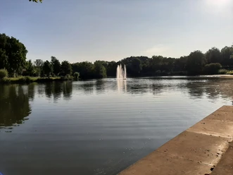 Gabelweiher Ruhiger Weiher mit Springbrunnen, umgeben von grünen Bäumen und Promenade, unter klarem Himmel.