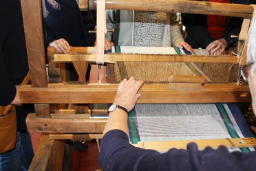 Webstuhl Ein traditioneller Webstuhl mit Holzstruktur, umgeben von Menschen, die das Weben beobachten oder helfen.