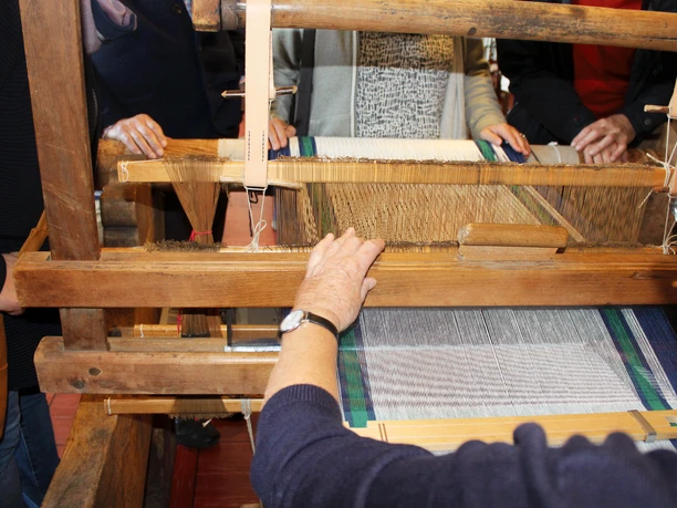 Webstuhl Ein traditioneller Webstuhl mit Holzstruktur, umgeben von Menschen, die das Weben beobachten oder helfen.