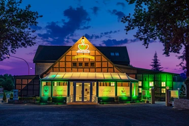 Dissen_Eventhaus Schamin_Aussenansicht_W_Schamin.jpg Eventhaus Schamin bei Sonnenuntergang, beleuchtet in Grün, mit Glasfassade und Fachwerkstruktur.