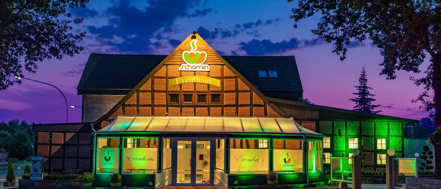 Dissen_Eventhaus Schamin_Aussenansicht_W_Schamin.jpg Eventhaus Schamin bei Sonnenuntergang, beleuchtet in Grün, mit Glasfassade und Fachwerkstruktur.