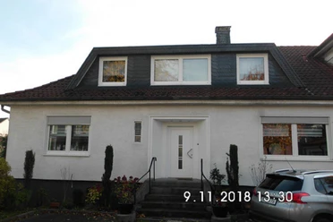 Ein weißes Einfamilienhaus mit schwarz gedecktem Dach, Treppe und seitlich parkendem Auto.