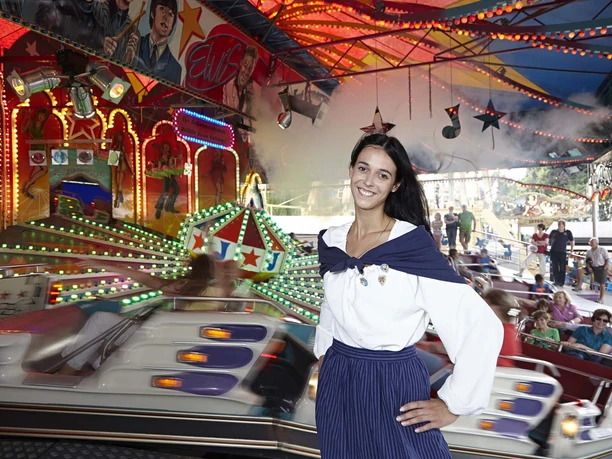 Frau in traditioneller Kleidung lächelt vor einem beleuchteten Fahrgeschäft auf dem Annentag in Brakel.