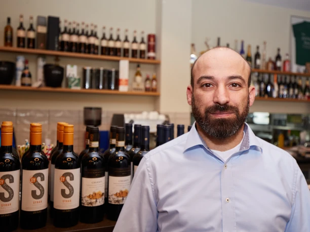 Ein freundlicher Mann steht vor einer Bar mit Regalen voller Weinflaschen und Spirituosen.