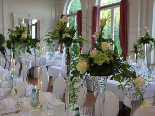 spiegelsaal-tischdeko-hochzeit.jpg Festlich gedeckter Hochzeitstisch im prächtigen Spiegelsaal mit eleganten Blumenarrangements aus weißen Rosen und Kerzen.