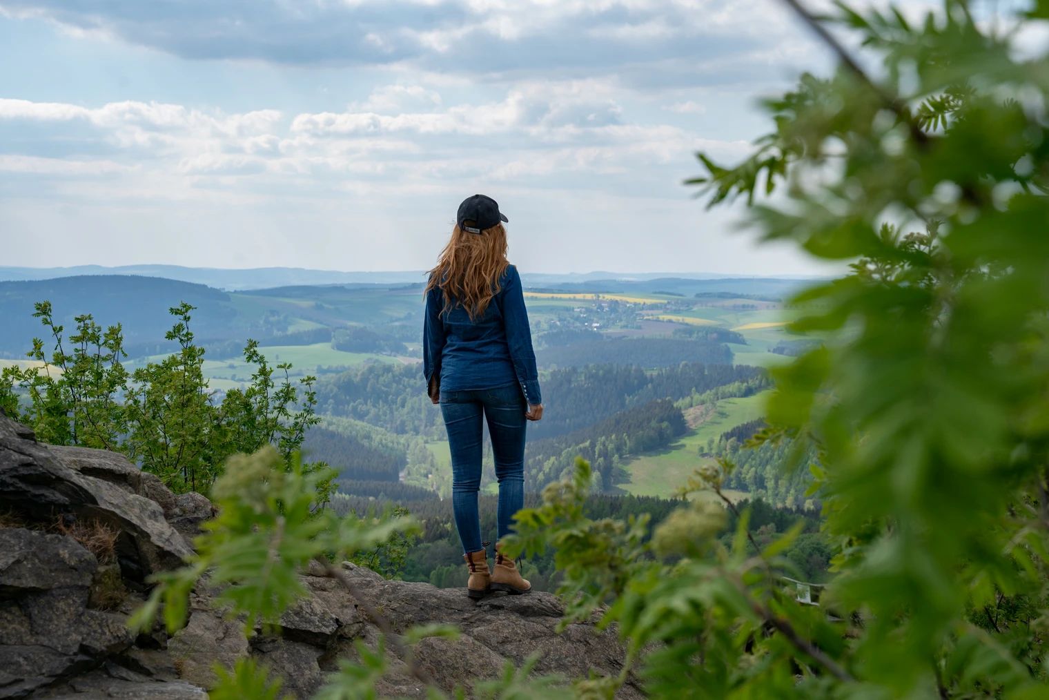 Am Schwartenberg (c) Thorsten Günthert (4).jpg Eine Frau steht auf einem Felsen und blickt über eine weite, grüne Hügellandschaft.