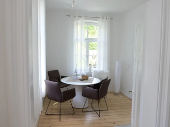 Kuesterhaus_3.JPG Heller Raum mit rundem Tisch, vier braunen Stühlen, Holzboden und einem Fenster mit transparenten Vorhängen.