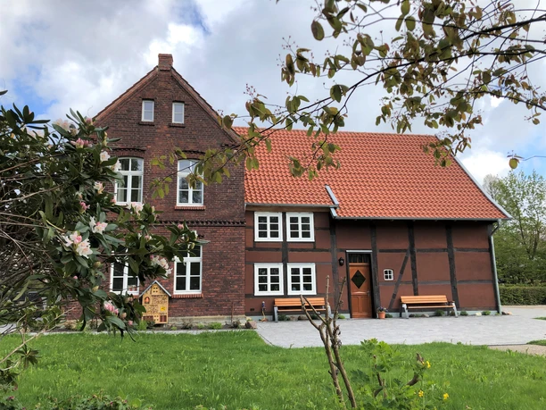 Backstein- und Fachwerkgebäude mit rotem Ziegeldach, umgeben von grüner Wiese und blühenden Bäumen.