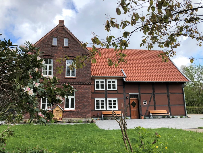 Backstein- und Fachwerkgebäude mit rotem Ziegeldach, umgeben von grüner Wiese und blühenden Bäumen.