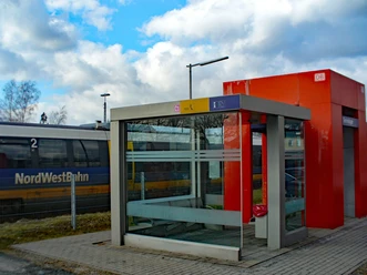 Ein kleiner Bahnhof mit moderner Wartehalle im Vordergrund, im Hintergrund ein NordWestBahn-Zug.