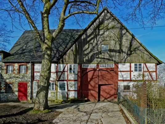 Fachwerkhaus mit Scheunentor und Baumschatten, umgeben von Natur; rustikaler ländlicher Eindruck.