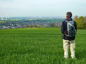 Mann im Grünen, blickt auf weite Landschaft mit Feldern, Ortschaft und Windrädern im Hintergrund.