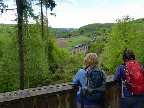 Blick von der Aussichtsplattform Dunetal auf den Kleinen Viadukt Zwei Wanderer blicken von einer hölzernen Plattform auf den Kleinen Viadukt im grünen Dunetal.