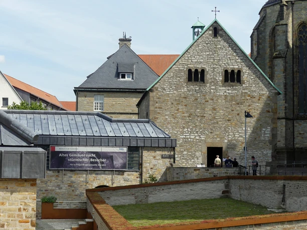 Steinbau mit Satteldach und quadratischem Turm, Flachdachgebäude und Bannerschrift im Vordergrund.
