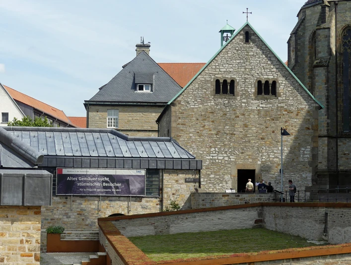 Steinbau mit Satteldach und quadratischem Turm, Flachdachgebäude und Bannerschrift im Vordergrund.