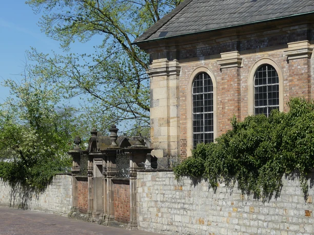 Die historische Alexiuskapelle mit alten Ziegelsteinen, flankiert von Bäumen und einem Steinzaun.