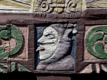 Holzrelief des Adam-und-Eva-Hauses zeigt ein ausdrucksstarkes Gesicht zwischen grünen Ornamenten.