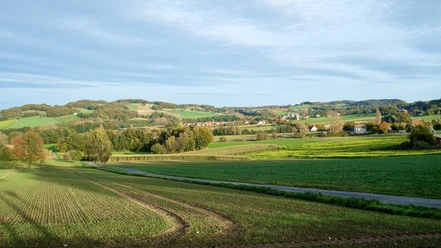 Ausblick Bonstapel Weite Felder unter blauem Himmel, sanfte Hügel im Hintergrund, vereinzelte Bäume und Häuser.