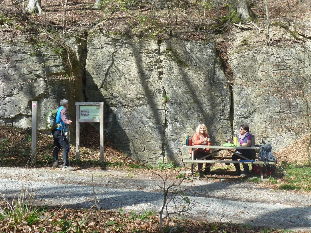 Ein Wanderer betrachtet eine Infotafel vor dem beeindruckenden Fels, daneben Menschen an einem Tisch.