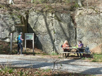 Steinbruch im Hanstein Ein Wanderer betrachtet eine Infotafel vor dem beeindruckenden Fels, daneben Menschen an einem Tisch.