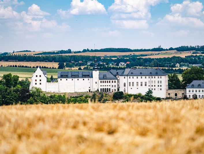 Schloss Wildenfels Schloss Wildenfels mit weißer Fassade und Schieferdach, umgeben von goldenen Feldern und Hügeln.Wildenfels Castle with its white façade and slate roof, surrounded by golden fields and hills.Zámek Wildenfels s bílou fasádou a břidlicovou střechou, obklopený zlatými poli a kopci.Zamek Wildenfels z białą fasadą i łupkowym dachem, otoczony złotymi polami i wzgórzami.Kasteel Wildenfels met zijn witte gevel en leistenen dak, omringd door gouden velden en heuvels.Il castello di Wildenfels con la sua facciata bianca e il tetto di ardesia, circondato da campi e colline dorate.