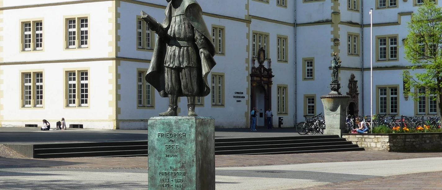 Bronzestatue Friedrich Spees, im Vordergrund eines historischen Schulgebäudes, mit Stufen.