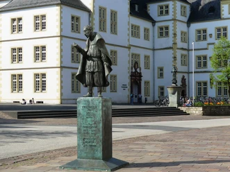 Spee-Statue vor dem Gymnasium Theodorianum Bronzestatue Friedrich Spees, im Vordergrund eines historischen Schulgebäudes, mit Stufen.
