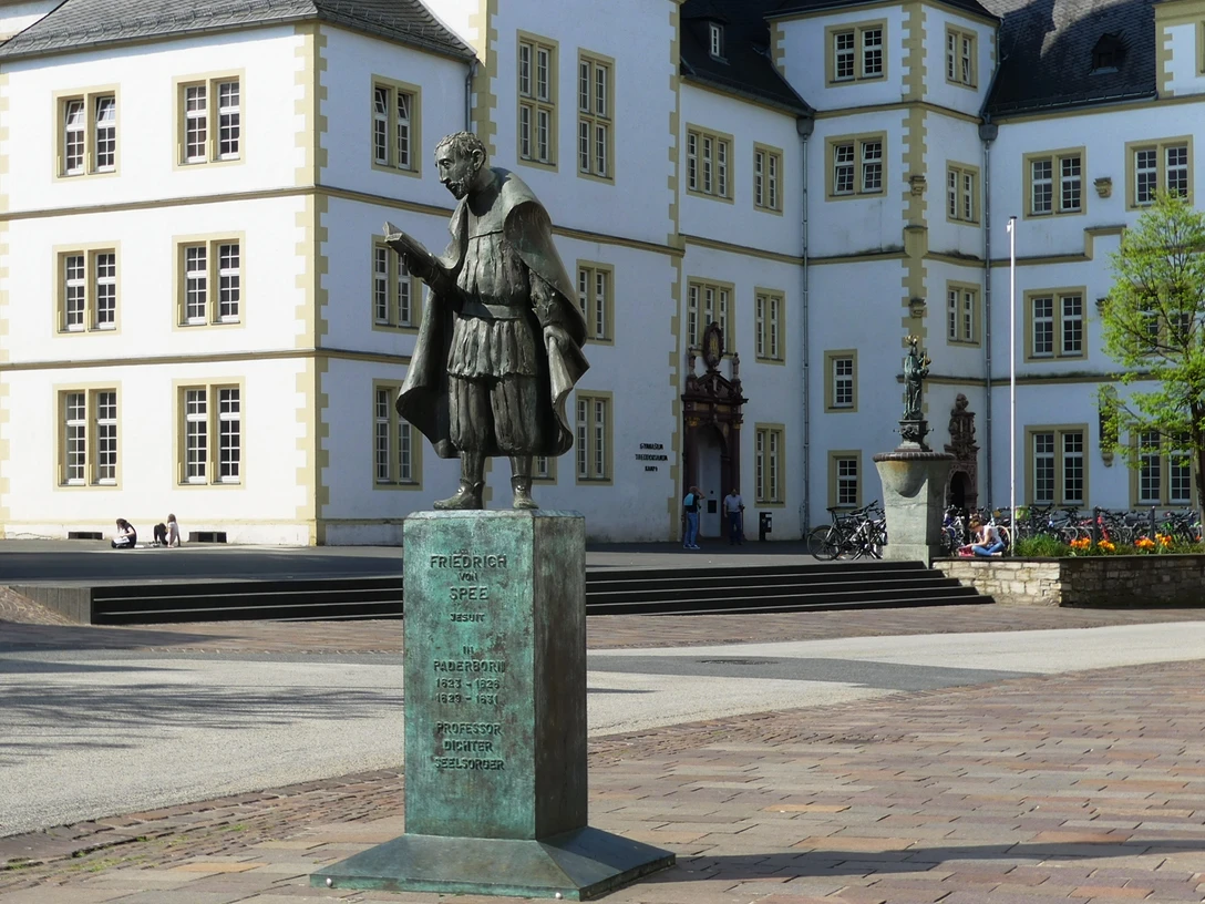 Spee-Statue vor dem Gymnasium Theodorianum Bronzestatue Friedrich Spees, im Vordergrund eines historischen Schulgebäudes, mit Stufen.