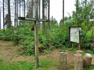 Gipfelkreuz auf bewaldetem Hügel nahe Paderborn, Wanderwegweiser und Infotafel inmitten von Bäumen.