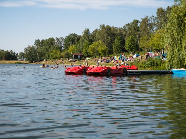Tretbootverleih Stausee Oberwald Rote Tretboote am ruhigen Ufer des Stausees mit Schwimmern und grüner Waldkulisse.