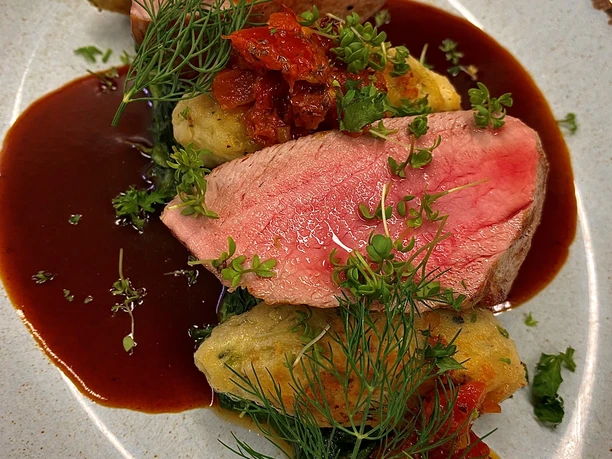 Ein kunstvoll angerichtetes Gericht mit rosa gebratenem Fleisch, Kräutern und Gemüse auf einem Teller.