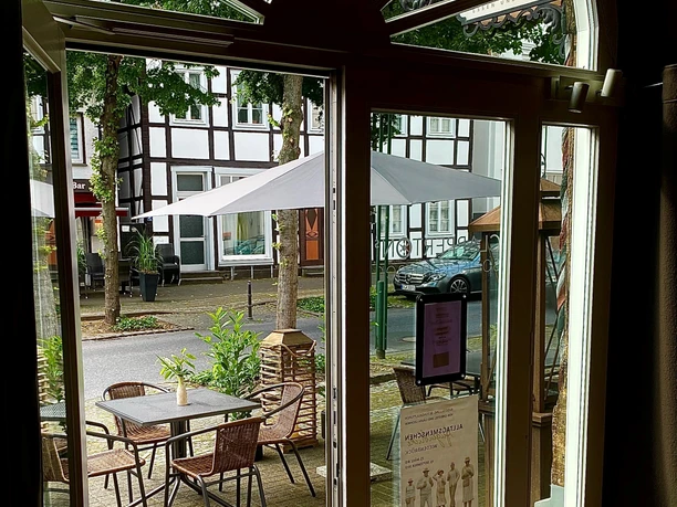 Blick durch ein Café-Fenster auf eine malerische, mit Bäumen gesäumte Terrasse mit Sonnenschirmen.