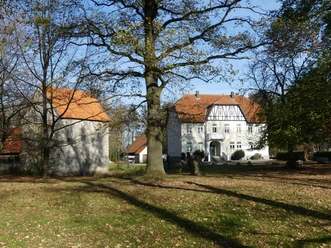 Ein weißes Fachwerkhaus mit roten Dächern steht inmitten von Bäumen und einem grünen Rasen.