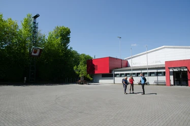 Mehrzweckhalle Dahl mit rotem Anbau, umgeben von Bäumen; drei Personen im Vordergrund auf dem Vorplatz.