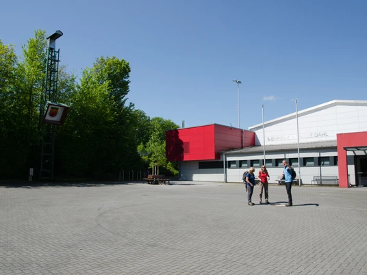 Mehrzweckhalle Dahl Mehrzweckhalle Dahl mit rotem Anbau, umgeben von Bäumen; drei Personen im Vordergrund auf dem Vorplatz.