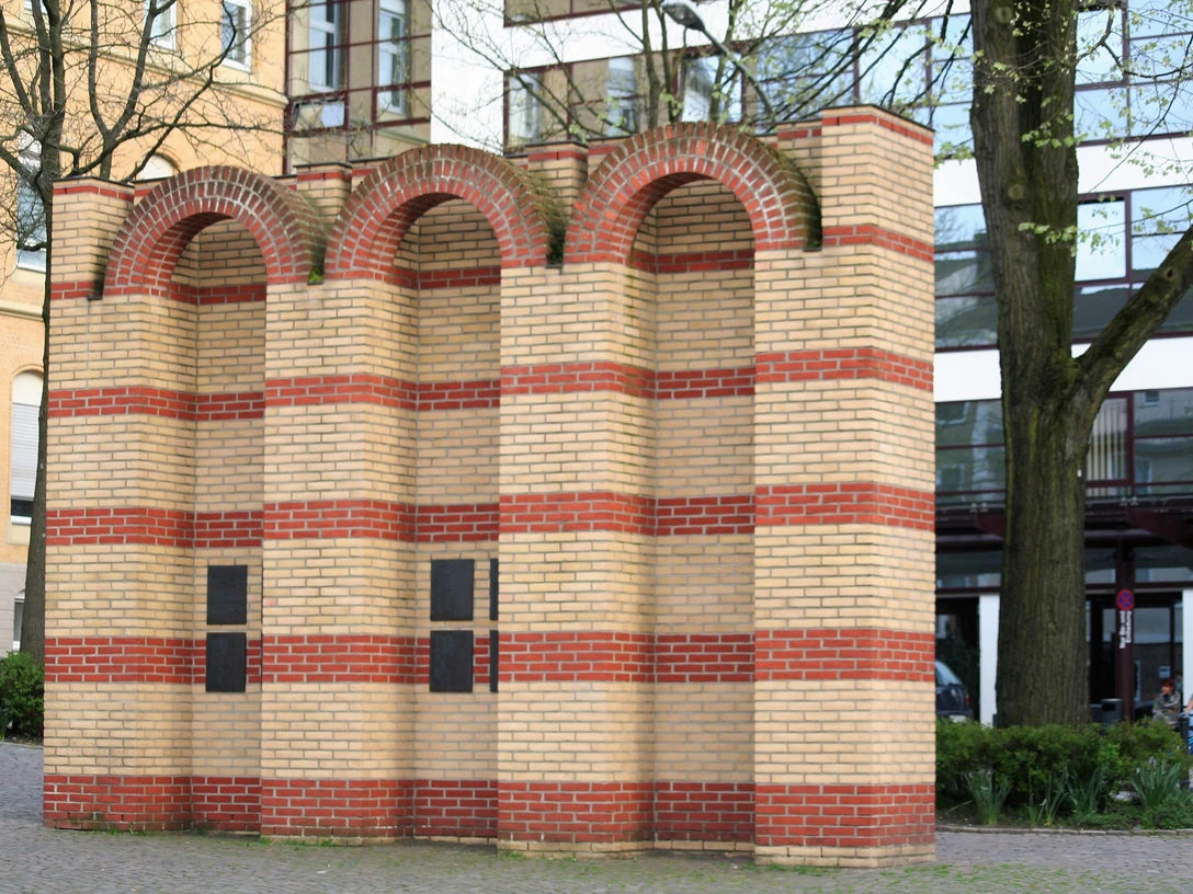 Drei segmentierte Backsteinbögen repräsentieren das jüdische Mahnmal, umgeben von moderner Architektur.