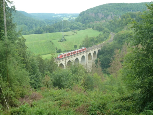 Blick von der Aussichtsplattform Dunetal auf den Kleinen Viadukt Panoramablick auf einen roten Zug, der über einen Viadukt in eine bewaldete, grüne Hügellandschaft fährt.
