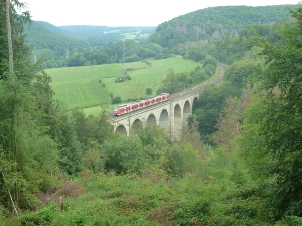 Blick von der Aussichtsplattform Dunetal auf den Kleinen Viadukt Panoramablick auf einen roten Zug, der über einen Viadukt in eine bewaldete, grüne Hügellandschaft fährt.
