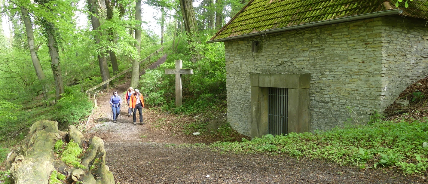 Zwei Wanderer nahe einem historischen, von Bäumen umgebenen Mausoleum mit einem großen Holzkreuz.
