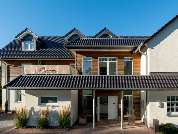 Albermann_1.jpg Gasthof mit moderner Fassade, blauen Dächern und großen Fenstern, umgeben von Pflanzen.