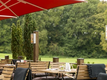 Holzterrasse mit gemütlichen Sitzgelegenheiten, roten Sonnenschirmen, umgeben von üppigem Grün.