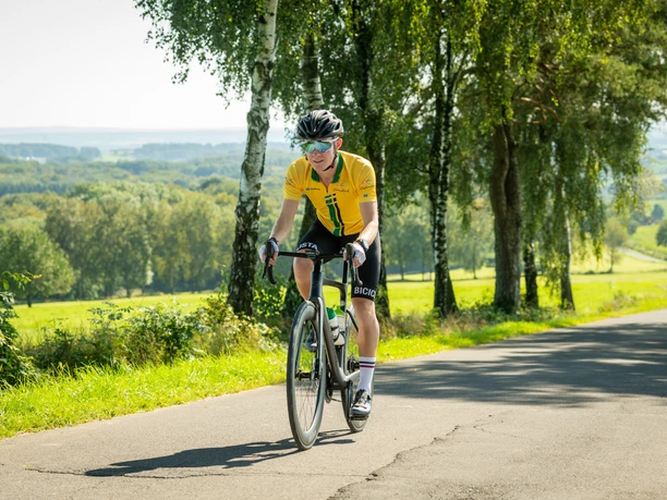 Rennrad Ein Radfahrer in gelbem Trikot fährt auf einer sonnigen, von Bäumen gesäumten Landstraße.
