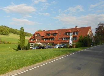 Ein traditionelles Gebäude mit rotem Satteldach vor einer grünen Wiesenlandschaft und blauen Himmel.