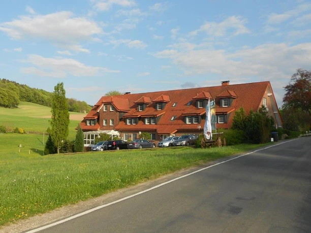 Ein traditionelles Gebäude mit rotem Satteldach vor einer grünen Wiesenlandschaft und blauen Himmel.