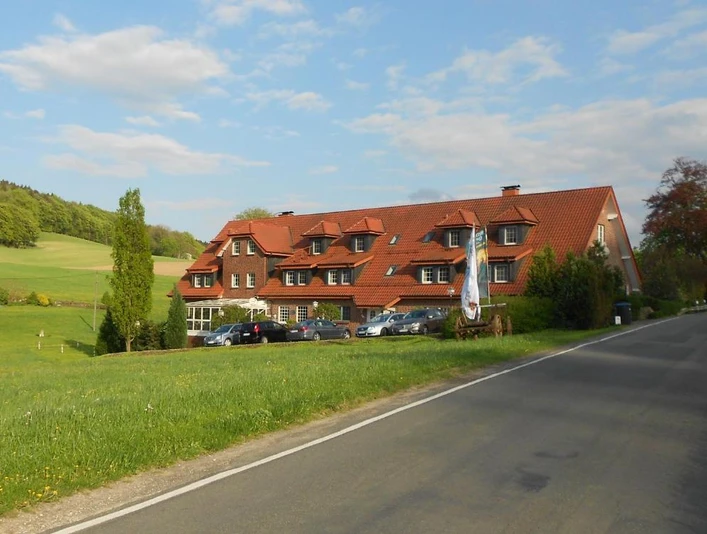 Außenansicht Ein traditionelles Gebäude mit rotem Satteldach vor einer grünen Wiesenlandschaft und blauen Himmel.