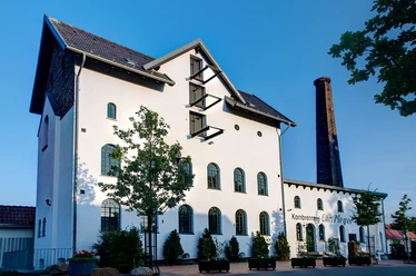 Ehemalige Brennerei mit weißer Fassade, Ziegeldach und hohem Schornstein vor blauem Himmel.