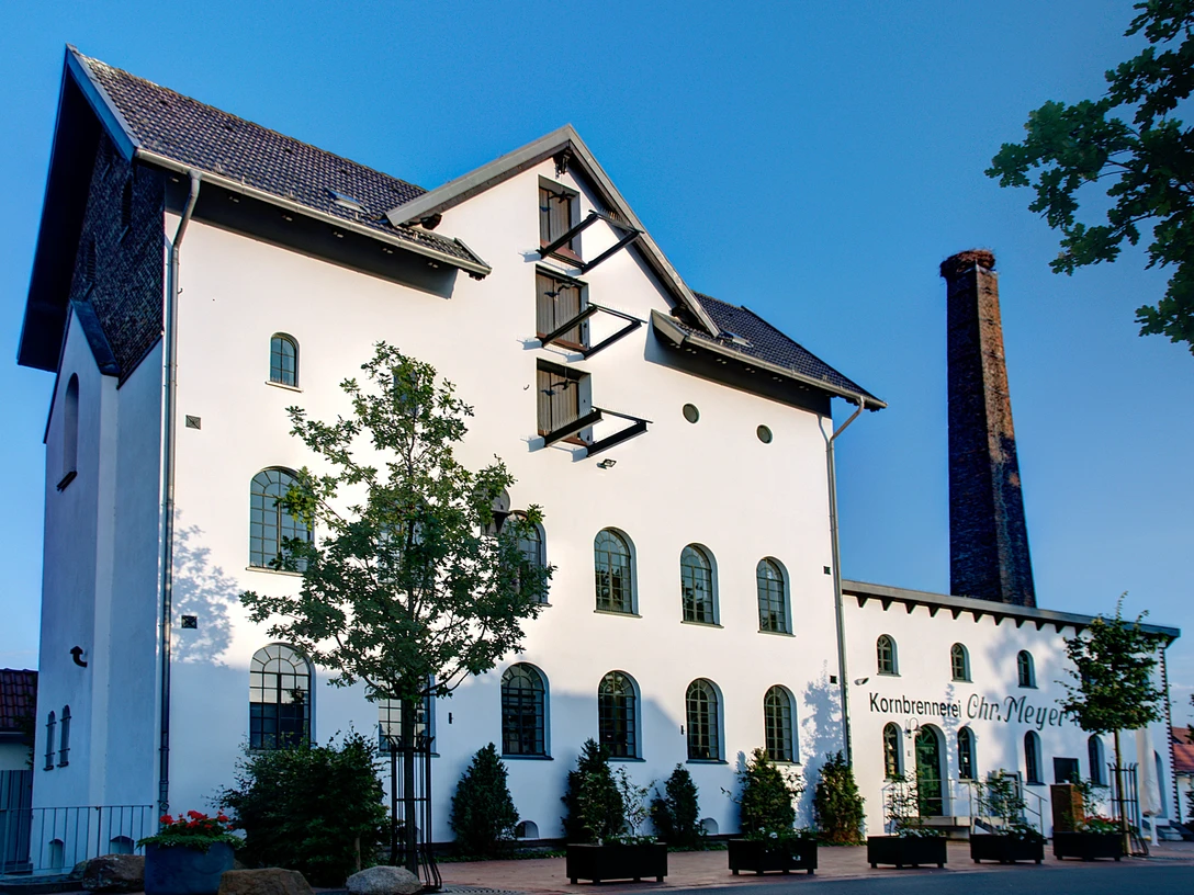 Alte Brennerei Hille Ehemalige Brennerei mit weißer Fassade, Ziegeldach und hohem Schornstein vor blauem Himmel.