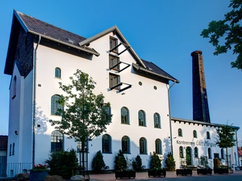 Alte Brennerei Hille Ehemalige Brennerei mit weißer Fassade, Ziegeldach und hohem Schornstein vor blauem Himmel.